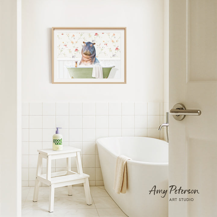A framed painting of a hippopotamus sitting in a bathtub hangs on a white tiled wall above a white bathtub in a bathroom.