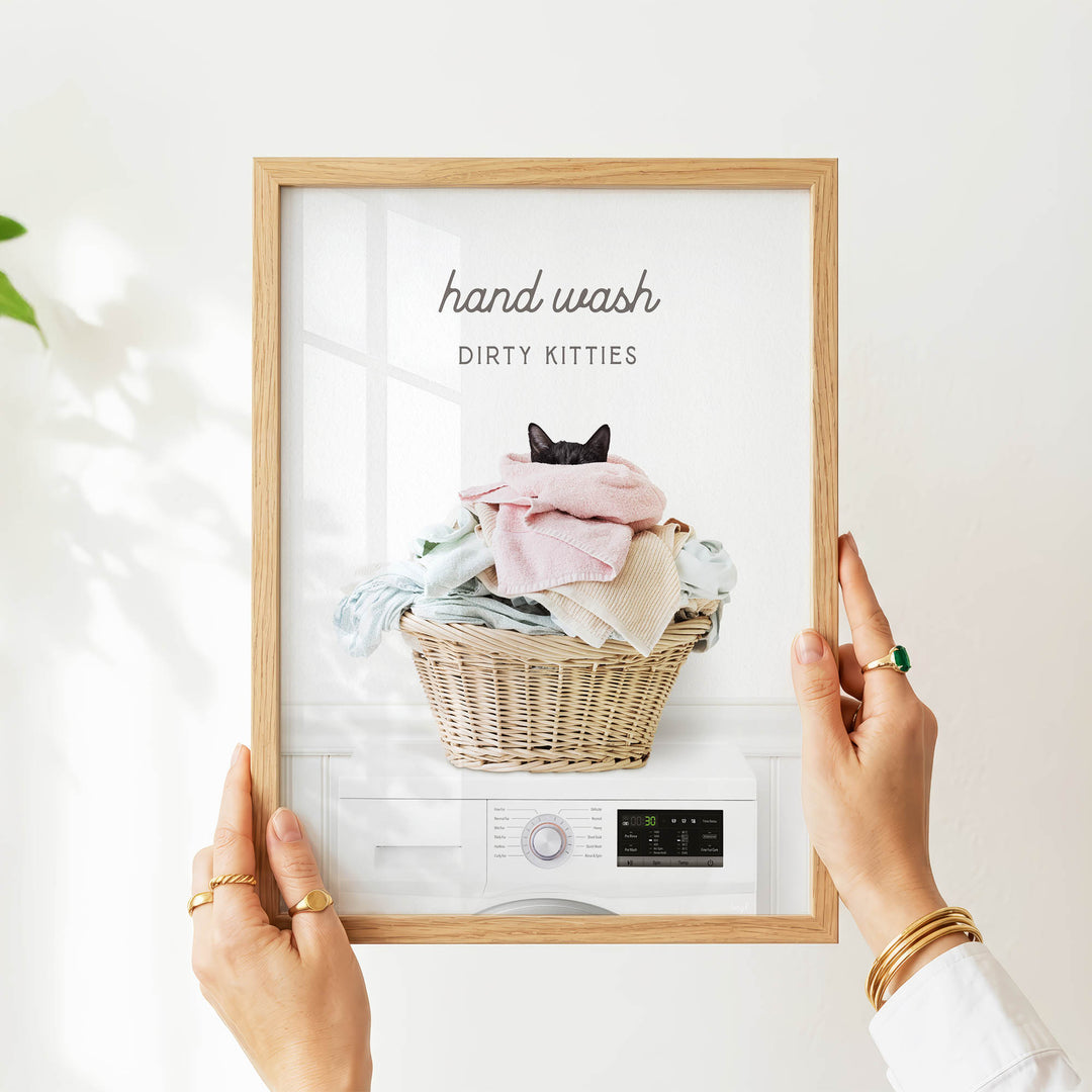 A person's hands are holding a framed picture of a cat sitting in a basket of laundry, with the text "hand wash dirty kitties" written above the image.