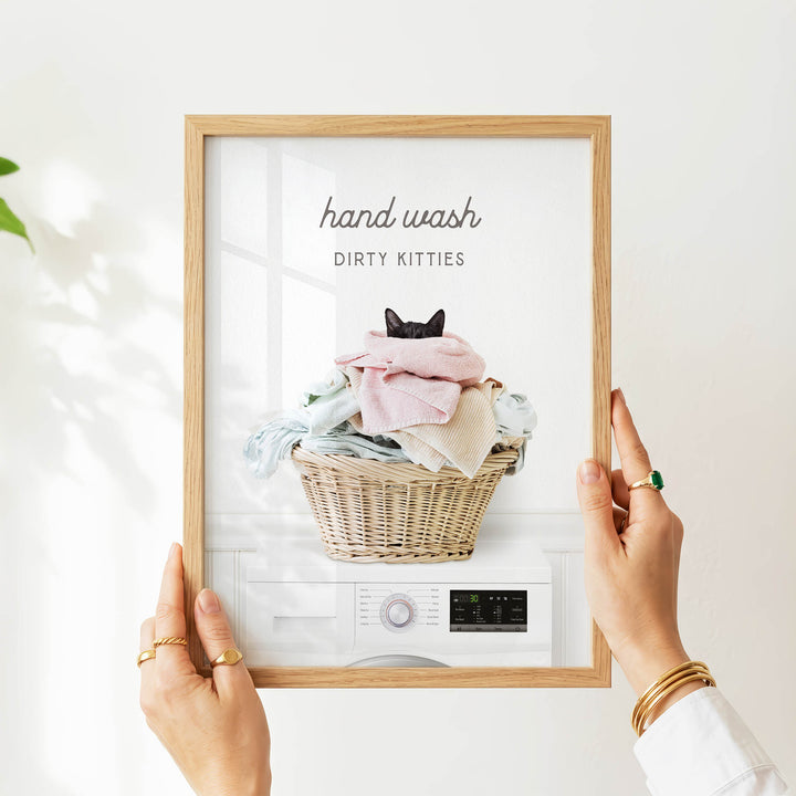 A person's hands are holding a framed picture of a cat sitting in a basket of laundry, with the text "hand wash dirty kitties" written above the image.