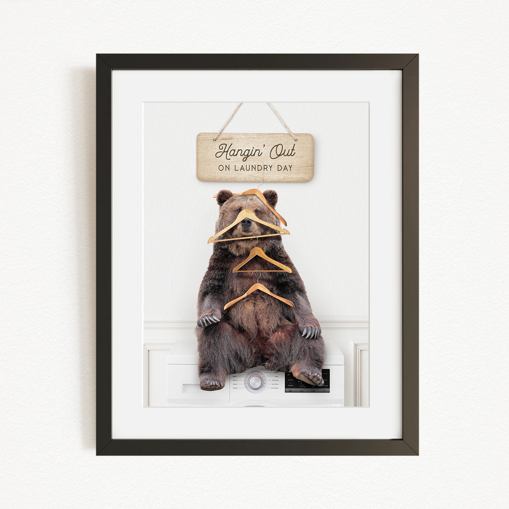 A framed image of a brown bear sitting on a white appliance, with a wooden sign above it that says "Hangin' Out on laundry day".