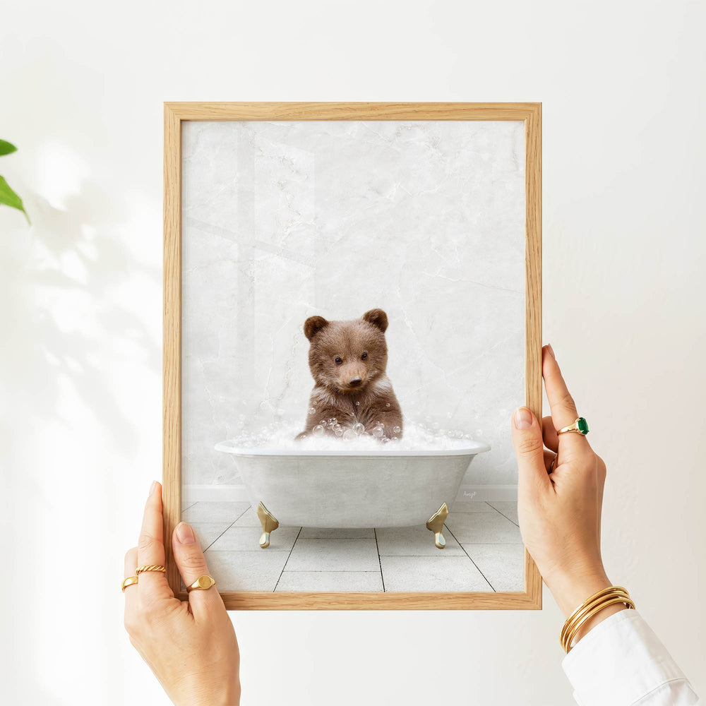 A person's hands are holding a framed picture of a baby bear sitting in a bathtub filled with water.
