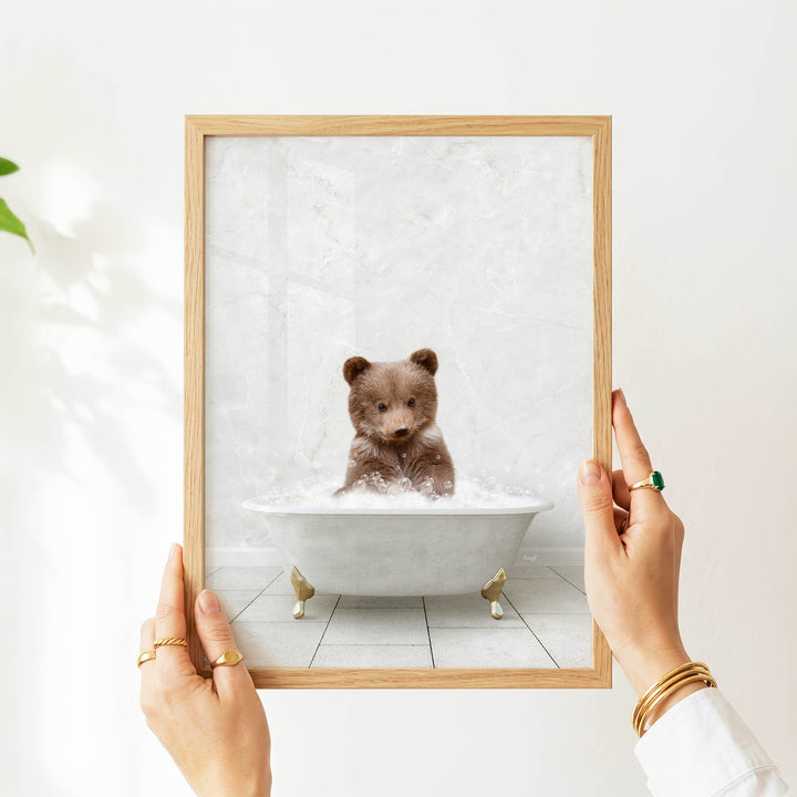 A person's hands are holding a framed picture of a baby bear sitting in a bathtub filled with water.
