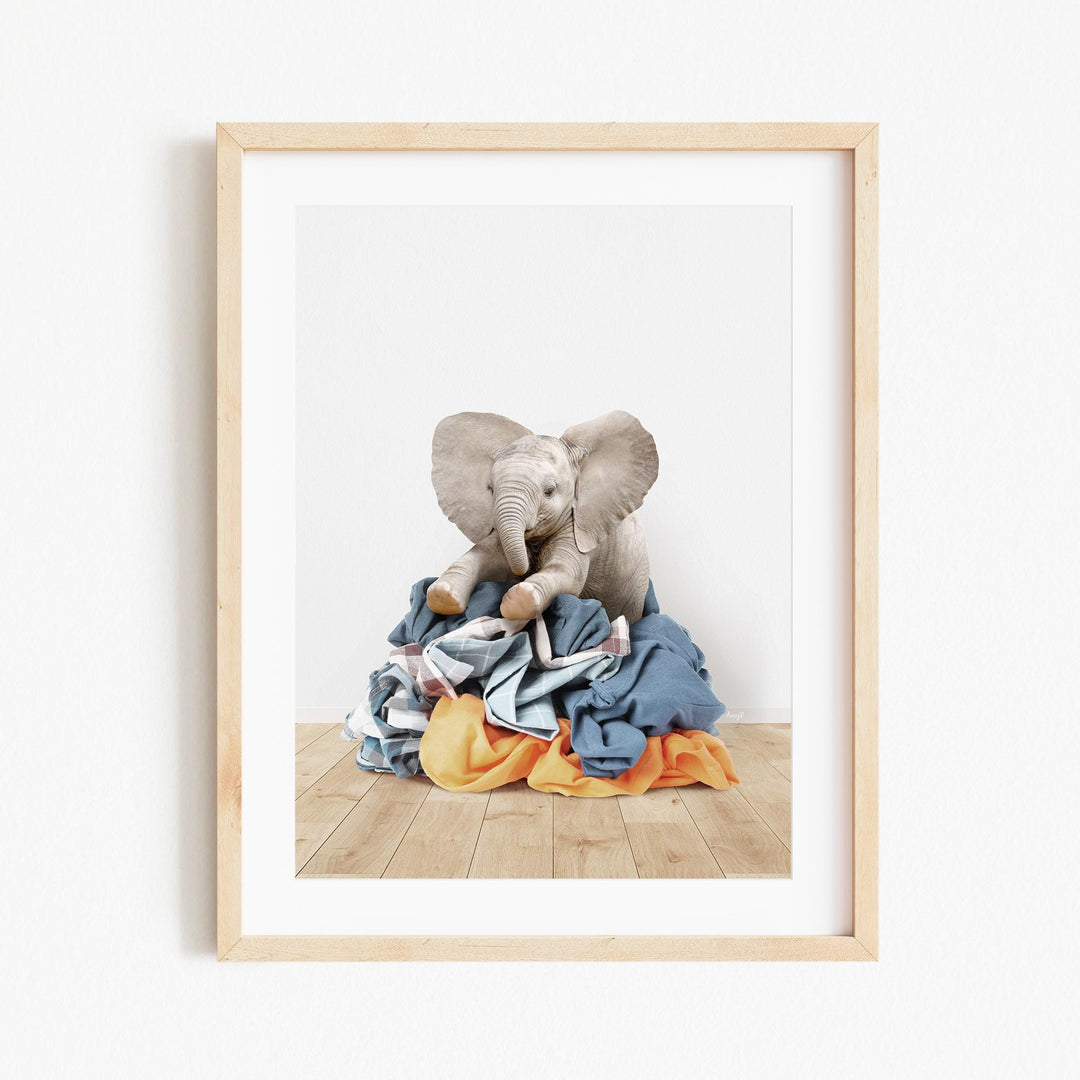 A gray elephant sits atop a pile of clothes on a wooden floor, creating a whimsical and playful scene.