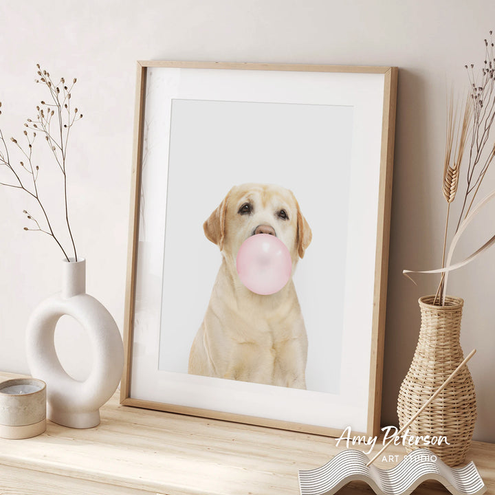 A framed photograph of a golden retriever dog blowing a bubble with a pink bubble gum bubble, accompanied by a wicker vase and a white ceramic vase on a wooden surface.