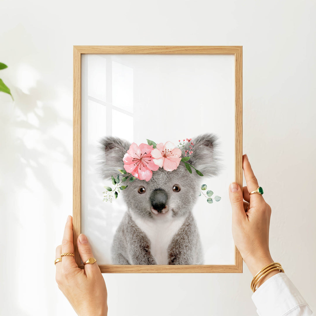 A person's hands are holding a wooden frame with a picture of a koala bear wearing a pink flower crown.