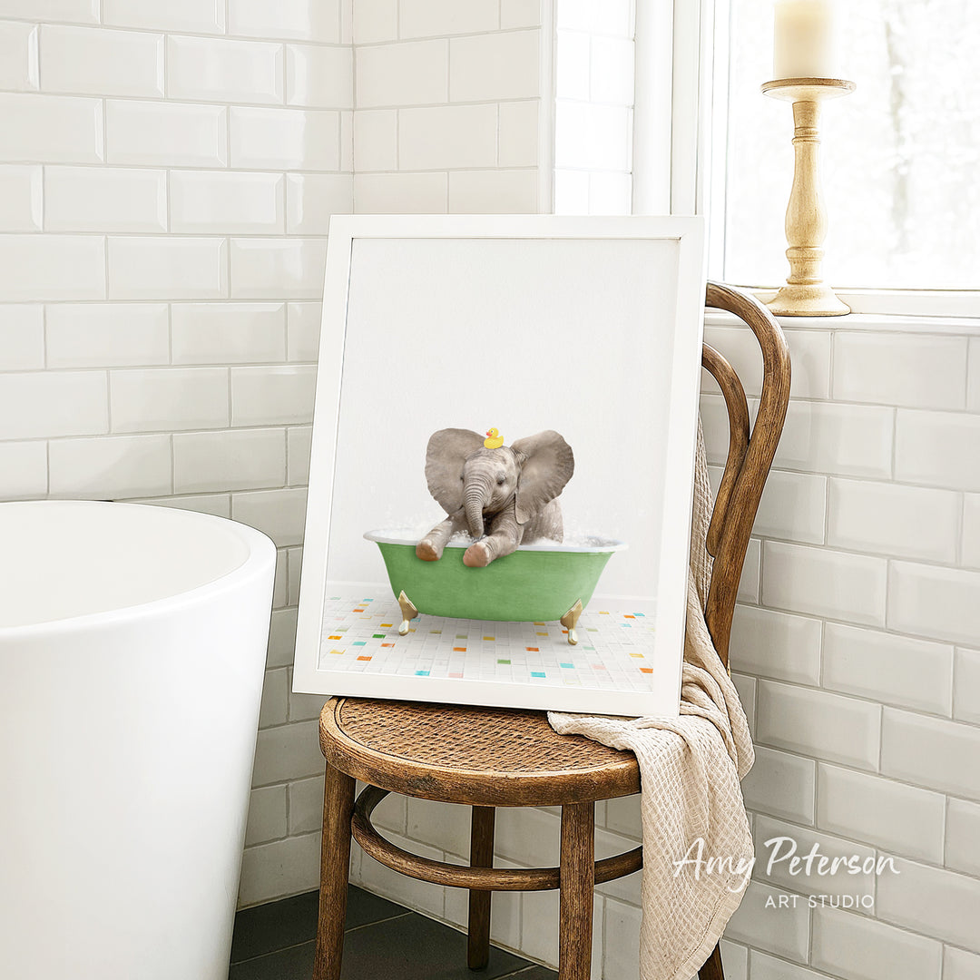 A framed image of a baby elephant sitting in a green bathtub, surrounded by colorful tiles, is displayed on a wooden chair in a bathroom setting.