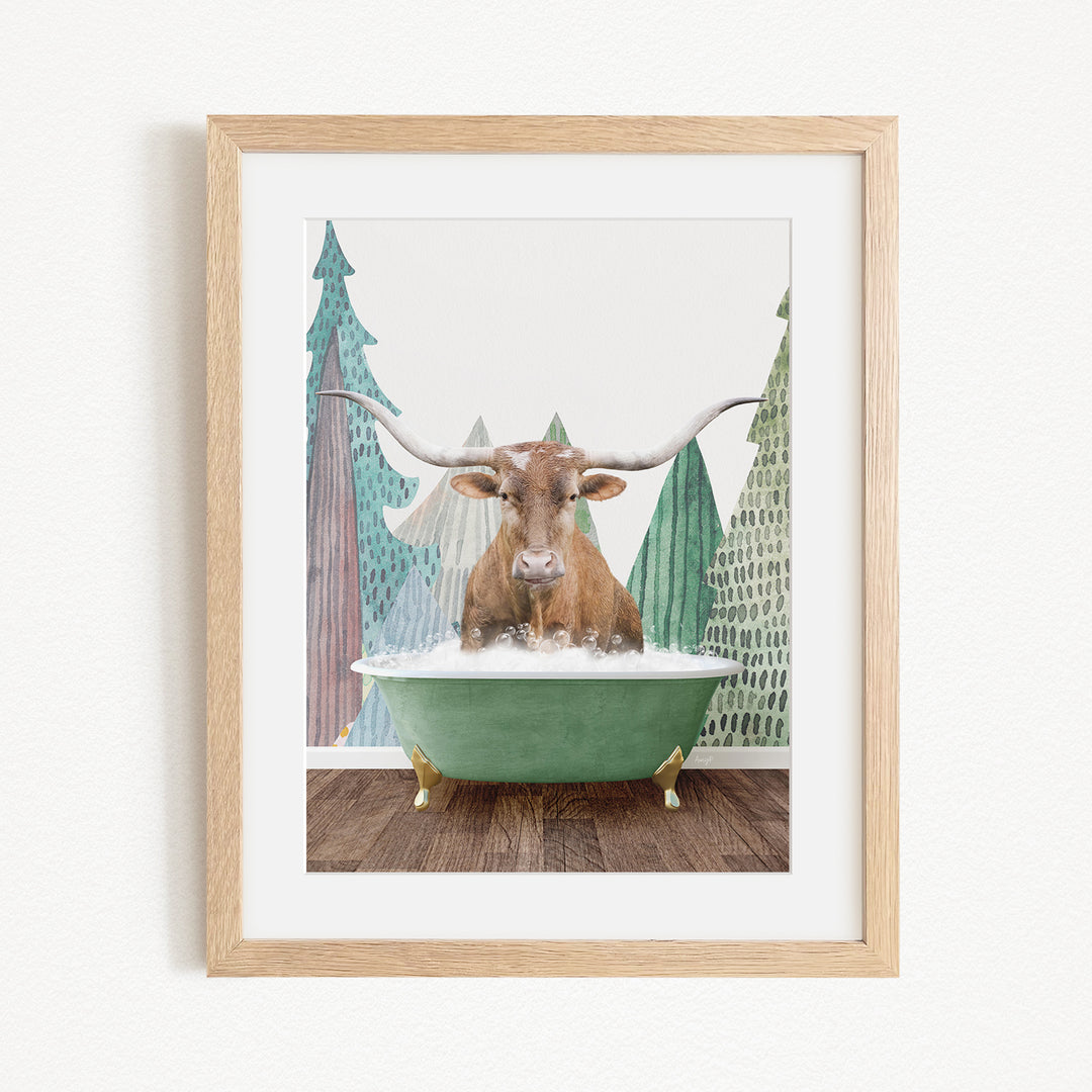 A framed print depicting a brown cow with large horns sitting in a green bathtub, surrounded by a forest-like background with pine trees and a snowy landscape.