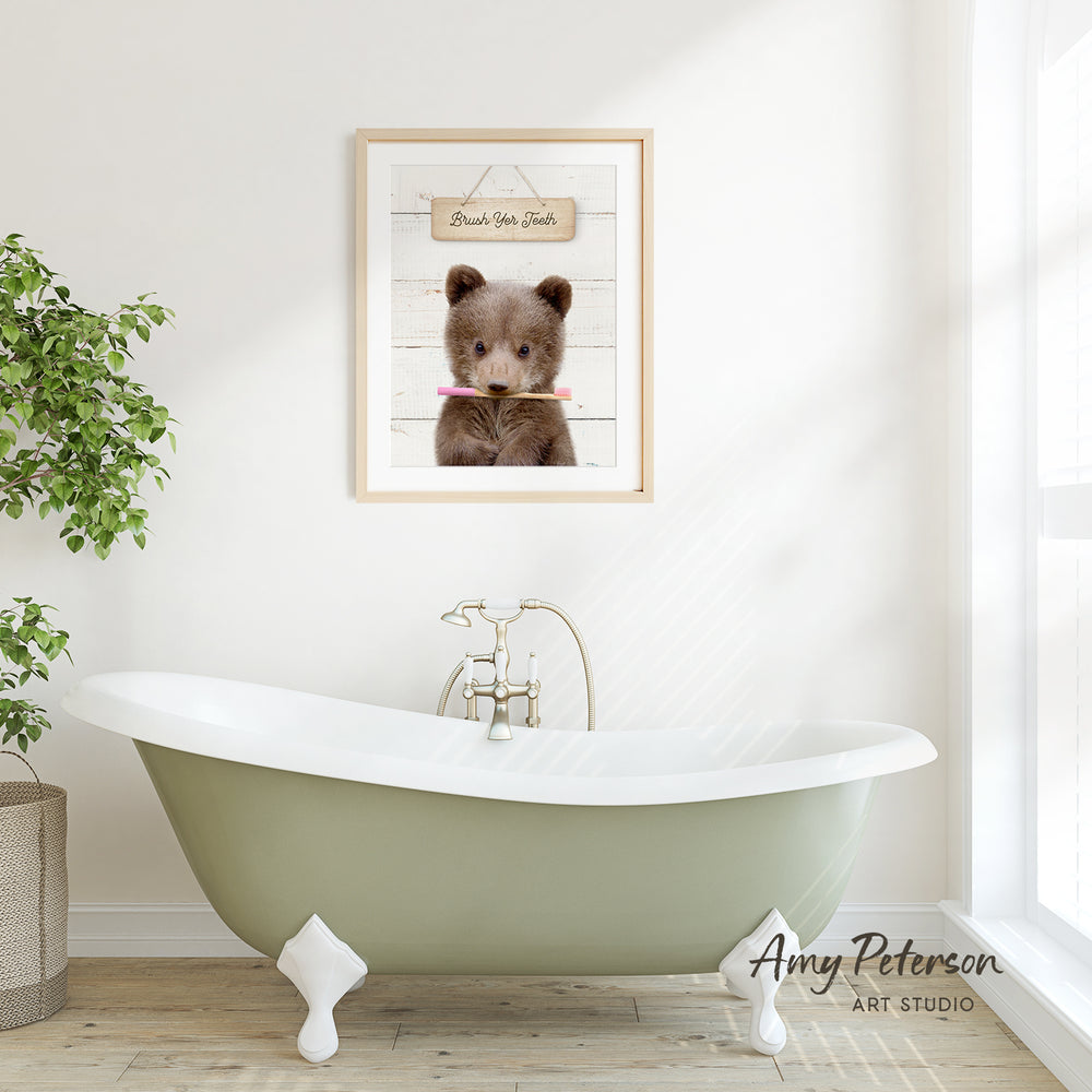 A framed image of a brown bear holding a toothbrush is displayed on a white wall above a green clawfoot bathtub in a bright, airy bathroom.