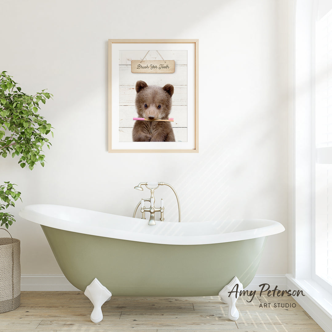 A framed image of a brown bear holding a toothbrush is displayed on a white wall above a green clawfoot bathtub in a bright, airy bathroom.
