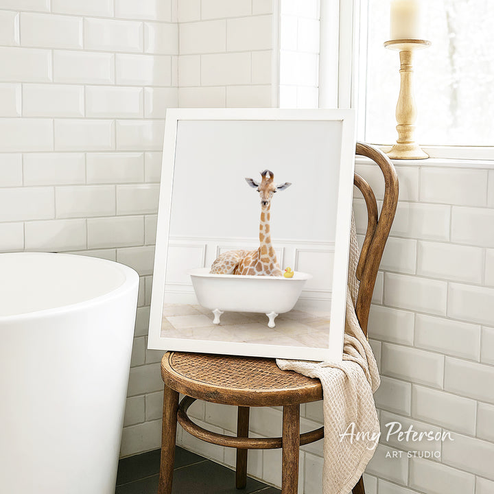 A framed image of a giraffe sitting in a white bathtub, with a yellow rubber duck floating in the water.