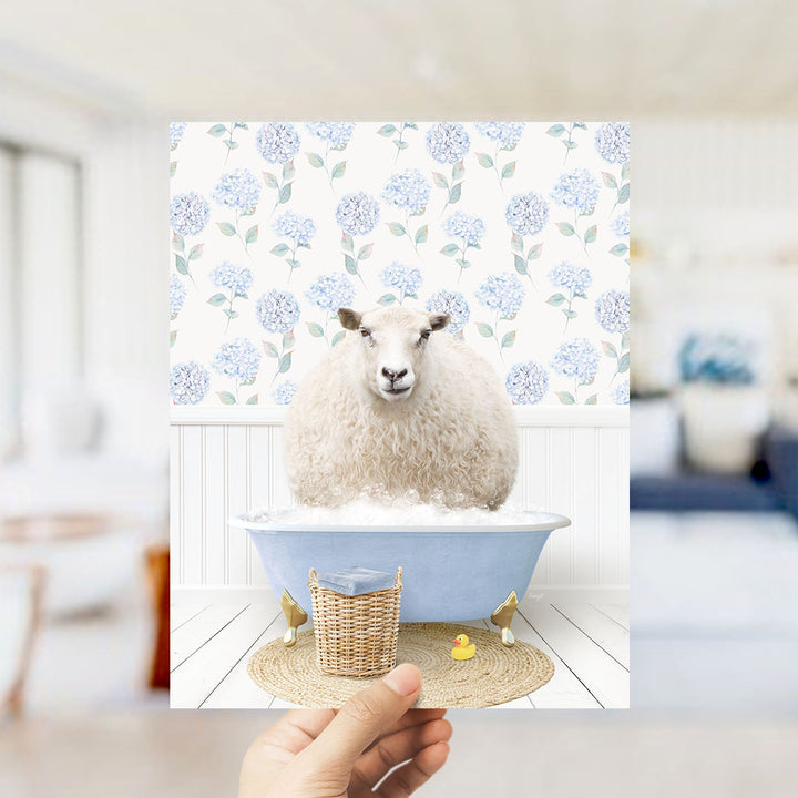 A hand holds up a framed image of a sheep sitting in a blue bathtub, surrounded by a wicker basket and a yellow rubber duck.