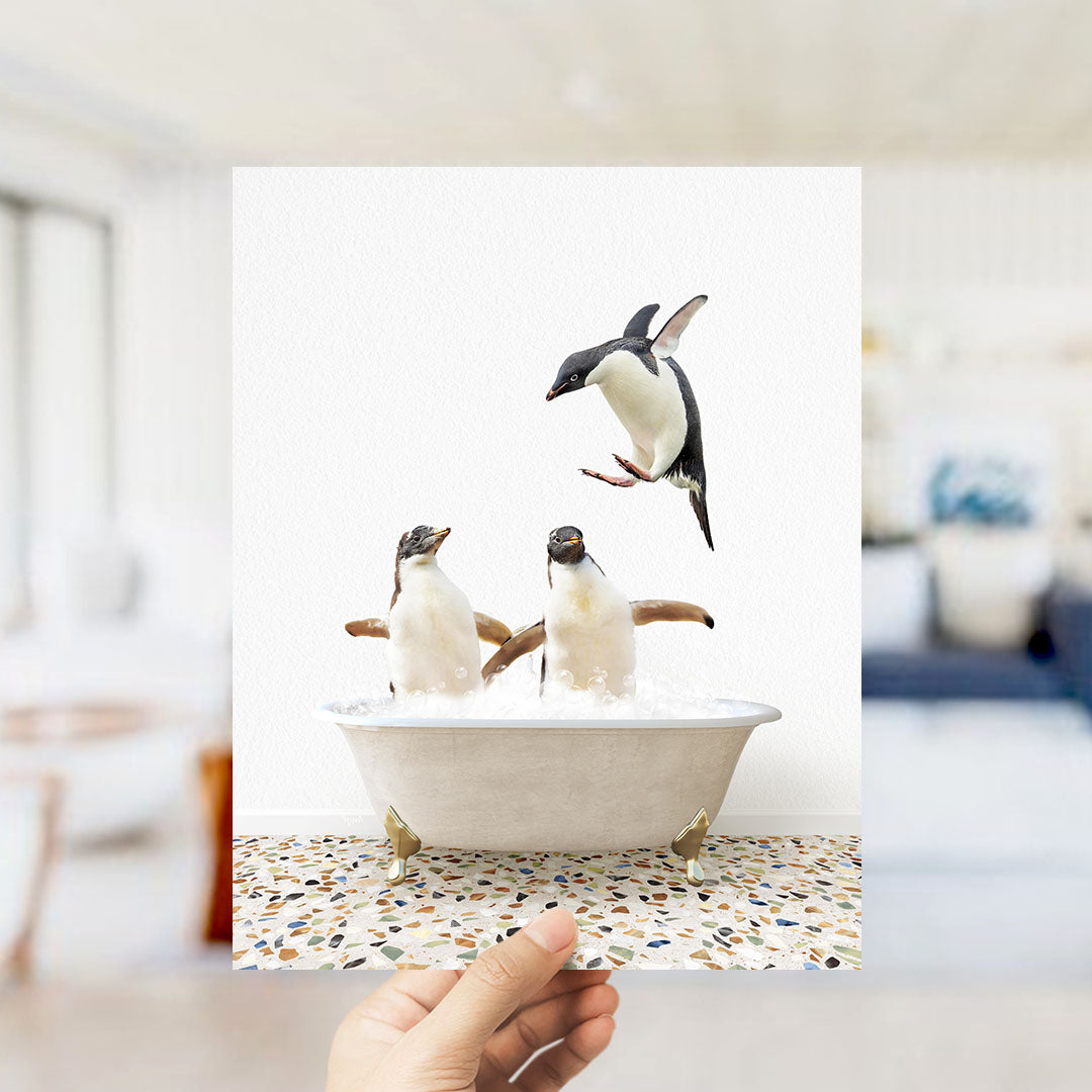 A hand holds up a framed photograph of two penguins standing in a bathtub filled with water, with one penguin flying out of the tub.