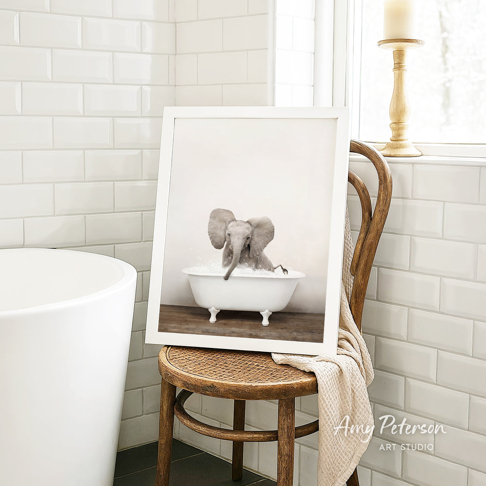 A framed photograph of an elephant sitting in a bathtub is displayed on a wooden chair in a bathroom setting.