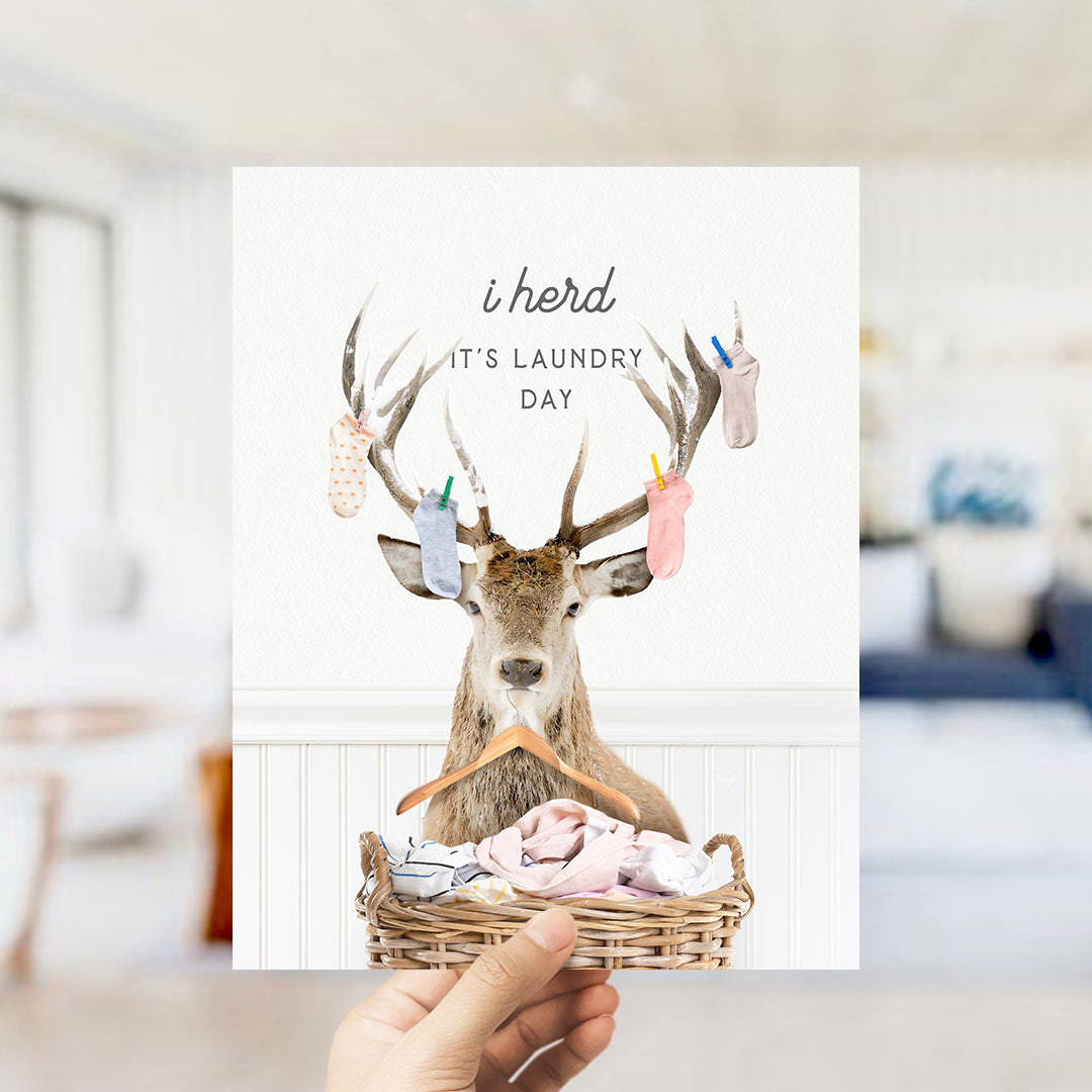 A hand holding a card with a deer wearing clothes and standing in a basket of laundry, with the text "It's laundry day" written above it.