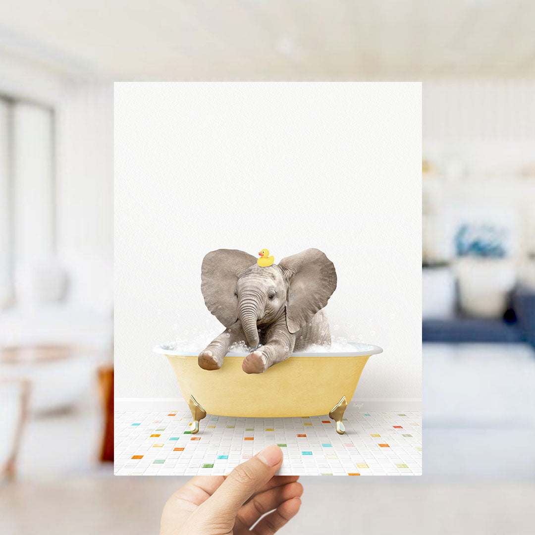 A hand holds up a framed image of a baby elephant sitting in a bathtub, with a yellow tub and colorful tiles on the floor.