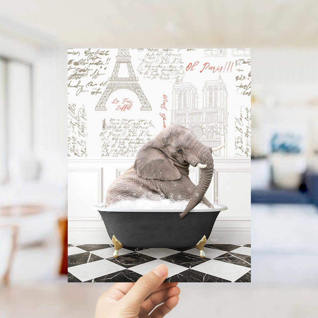 A hand holds up a printed image featuring an elephant sitting in a bathtub filled with bubbles, with the Eiffel Tower and other Parisian landmarks visible in the background.