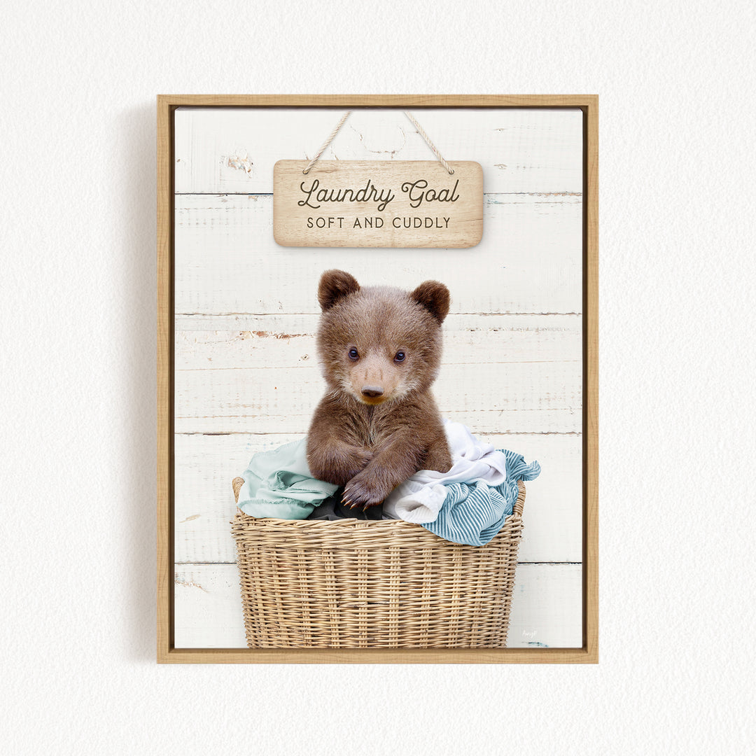 A cute brown bear sitting in a wicker laundry basket, with a wooden sign above that says "Laundry Goal Soft and Cuddly".