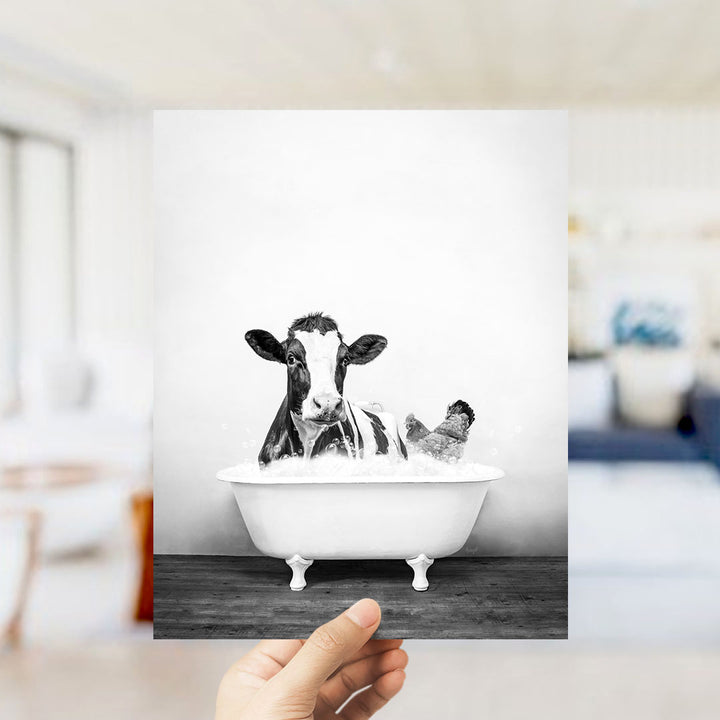 A black and white photograph of a cow sitting in a bathtub, with a person's hand holding the photo up to the camera.