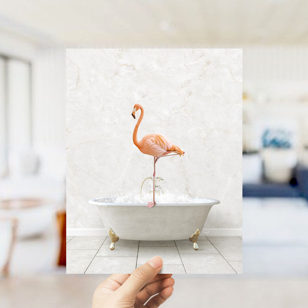 A hand holds up a framed image of a pink flamingo standing in a white bathtub filled with bubbles.