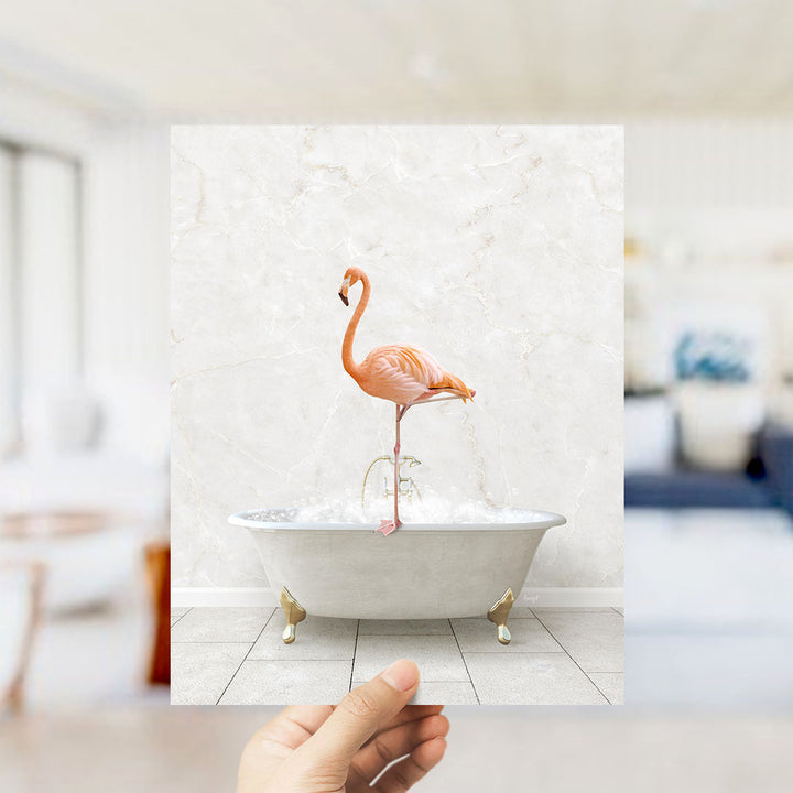 A hand holds up a framed image of a pink flamingo standing in a white bathtub filled with bubbles.