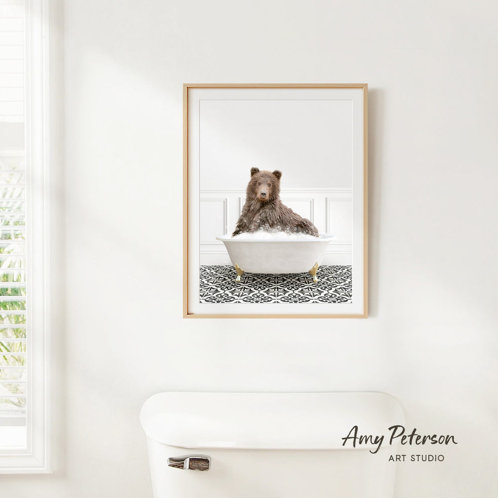 A framed image of a brown bear sitting in a white bathtub, with a patterned tile floor beneath it, is hanging on a white wall.