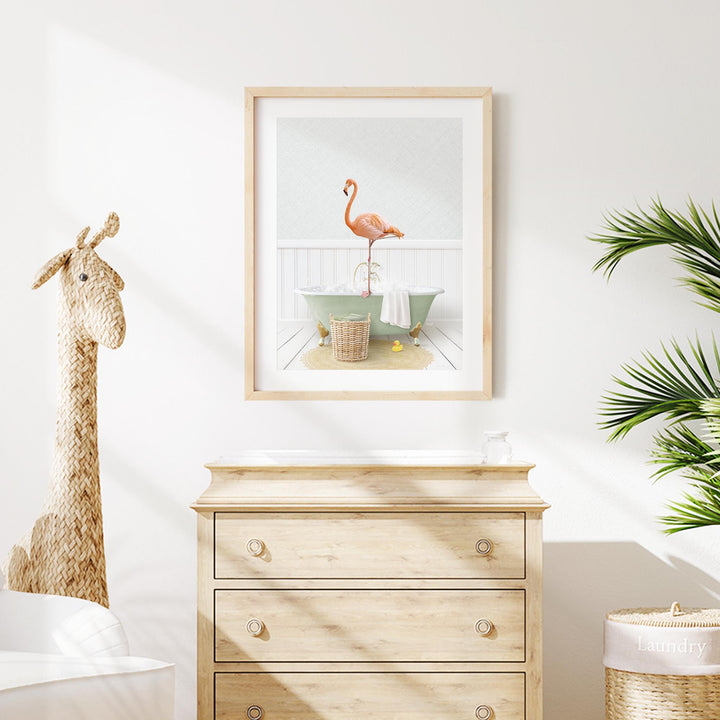 a framed print of a pink flamingo standing on a bathtub, with a wooden dresser below it and a stuffed giraffe toy to the left.
