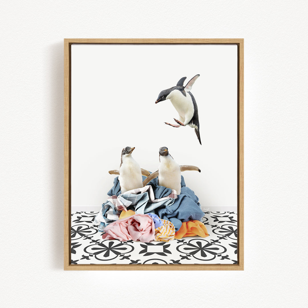 A framed photograph depicting two penguins standing on a pile of clothes, with one penguin flying in the air above the other.
