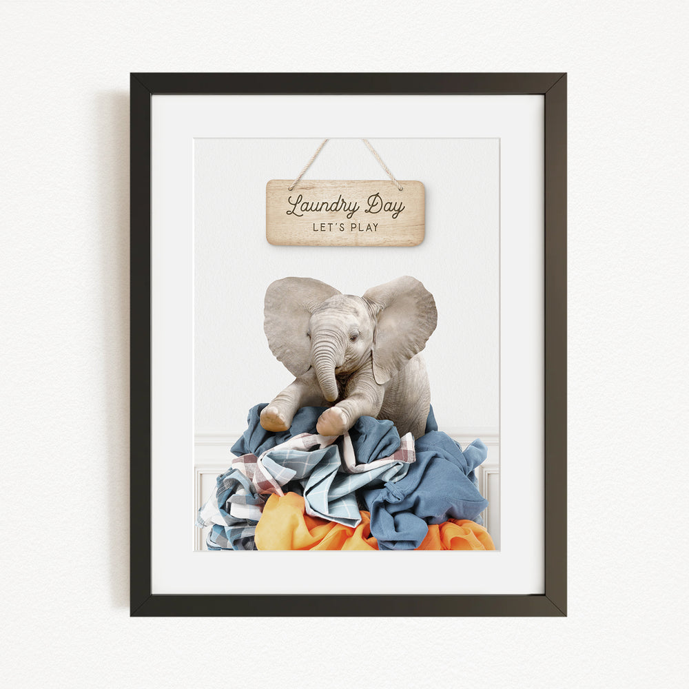 A framed image of a gray elephant sitting atop a pile of clothes, with a wooden sign above that says "Laundry Day Let's Play".