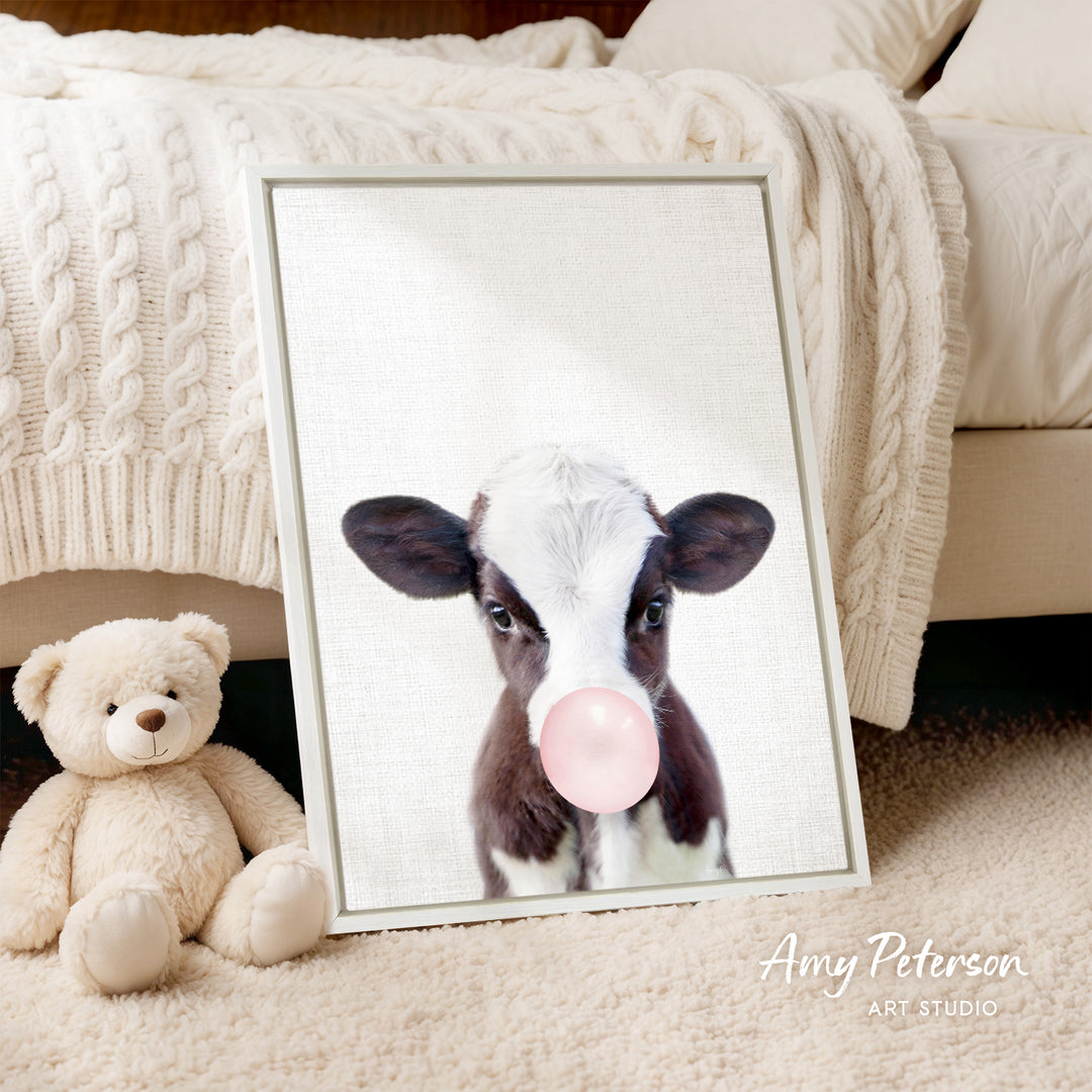 A framed image of a cow blowing a bubble gum bubble is displayed on a bed, accompanied by a teddy bear.