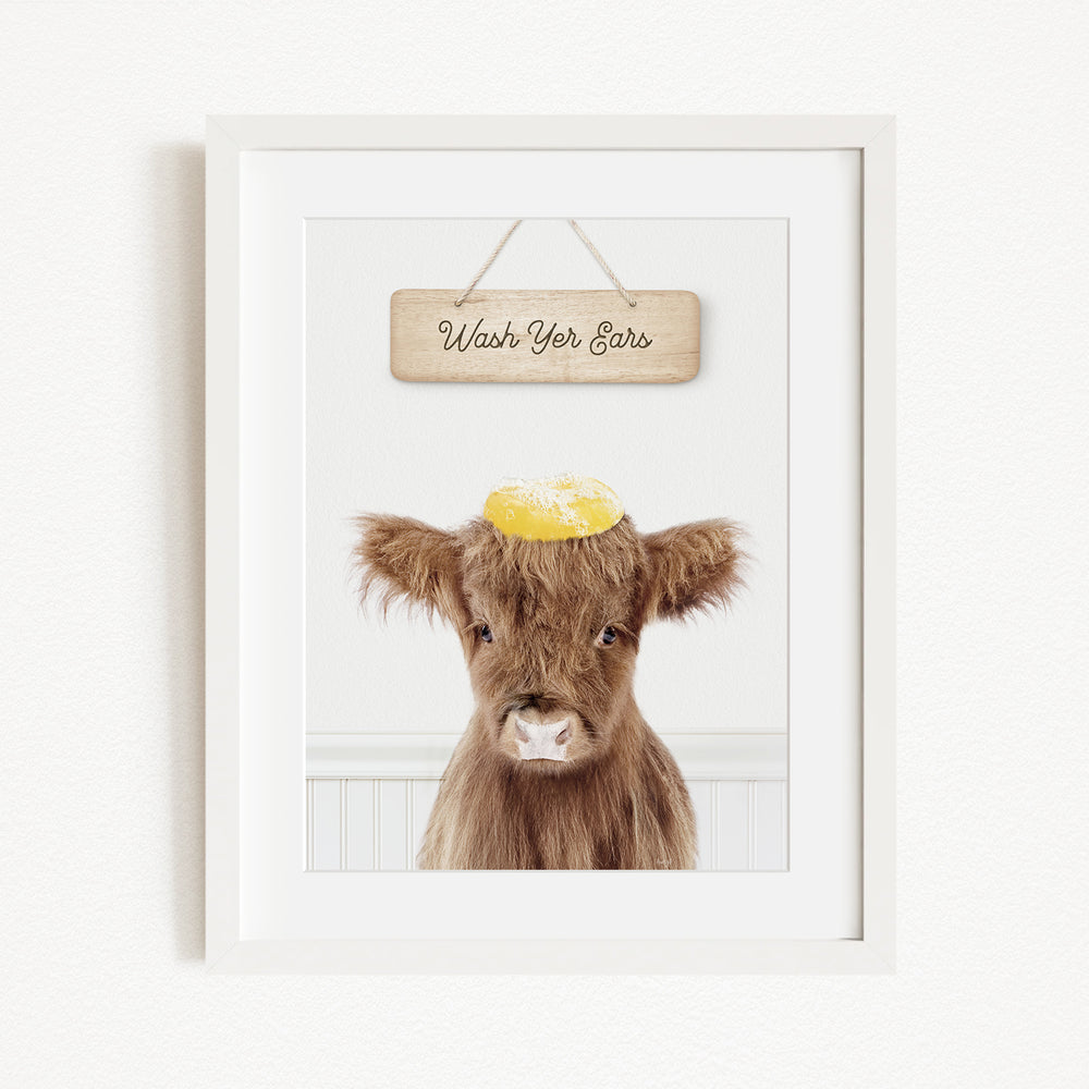 A framed image of a brown cow with a yellow object on its head, accompanied by a wooden sign that says "Wash Your Ears".