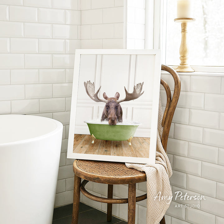 A framed image of a moose sitting in a green bathtub, with its antlers raised, is displayed on a wooden chair in a bathroom setting.