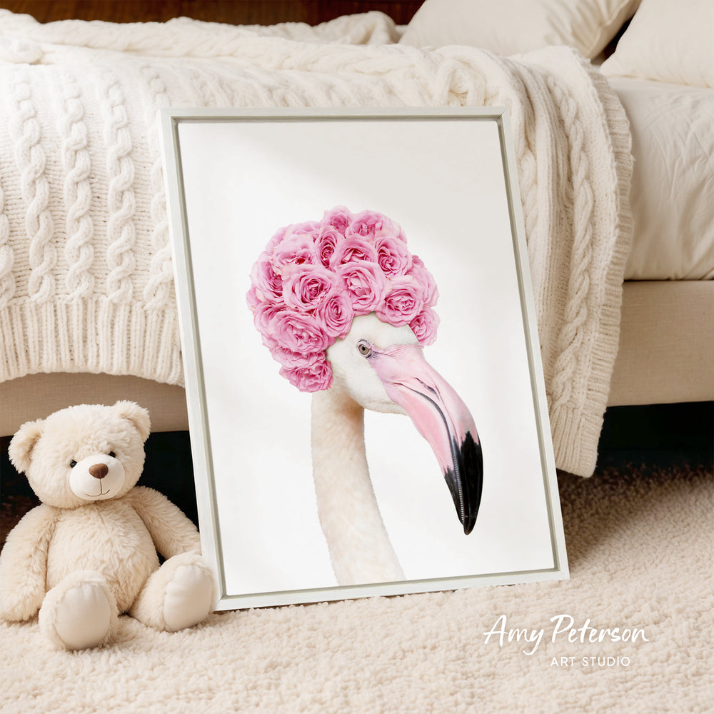 A framed image of a flamingo with a pink flower crown on its head, placed on a white bedspread and a white teddy bear.
