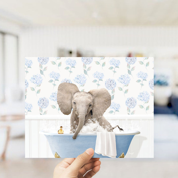 A hand holds up a framed image depicting a baby elephant sitting in a blue bathtub, surrounded by a floral pattern.