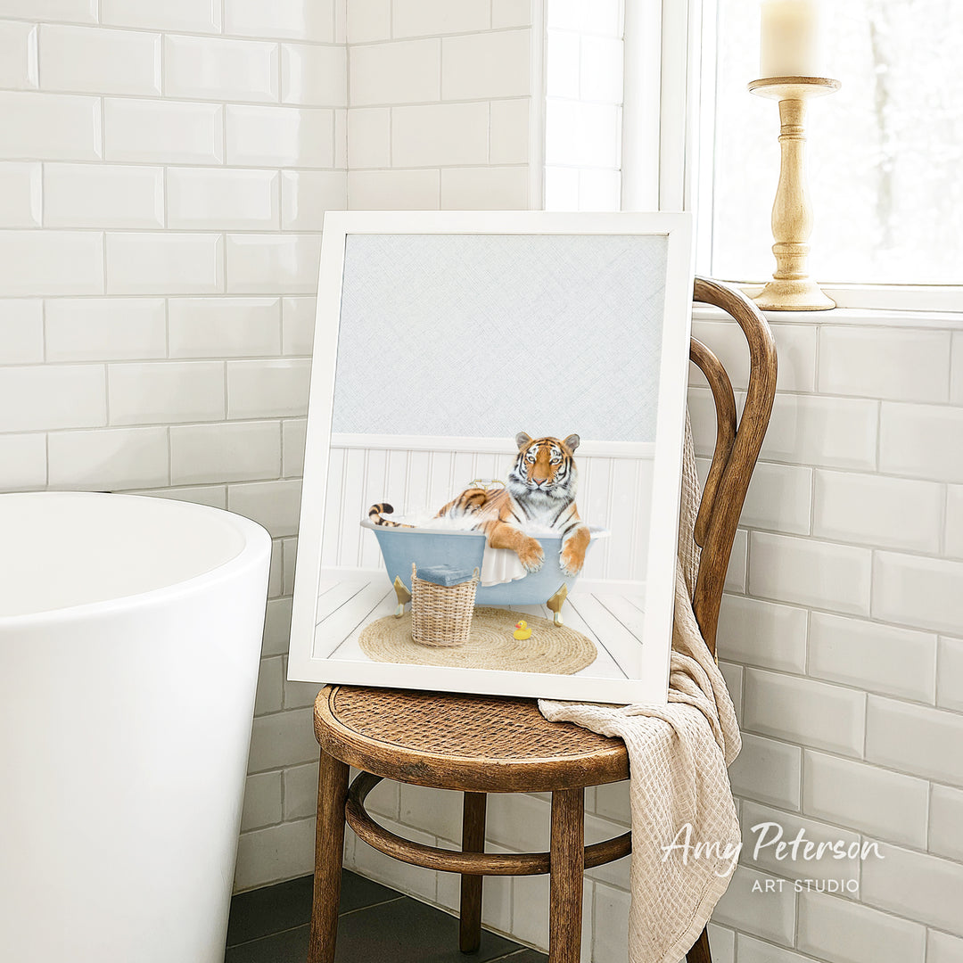 A framed image of a tiger sitting in a bathtub, surrounded by a basket of toys, is displayed on a wooden chair in a bathroom setting.