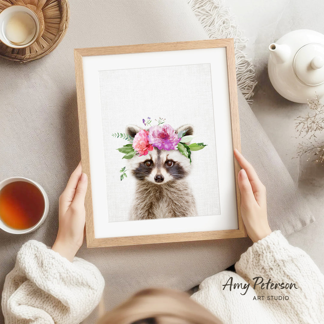 A person is holding a framed picture of a raccoon wearing a floral crown, with a teapot, a cup of tea, and a bowl of tea nearby.