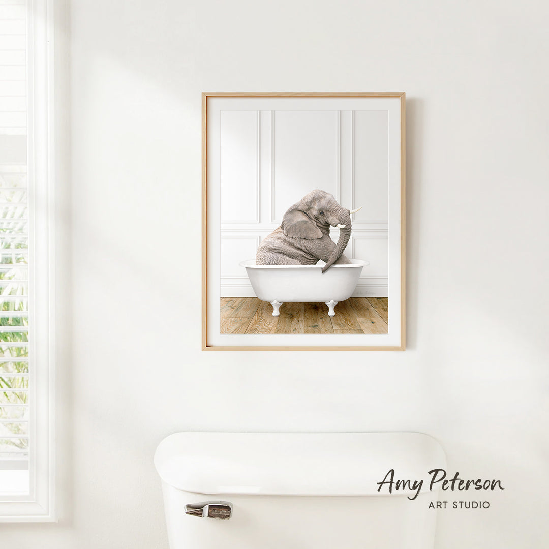 A framed image of an elephant sitting in a bathtub is hanging on a white wall.