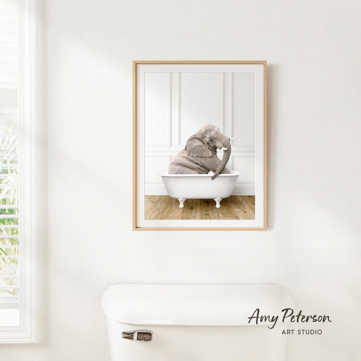 A framed image of an elephant sitting in a bathtub is hanging on a white wall.
