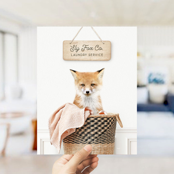 A hand holding a framed photograph of a small orange fox sitting in a woven basket, with a sign above it that reads "Laundry Service".