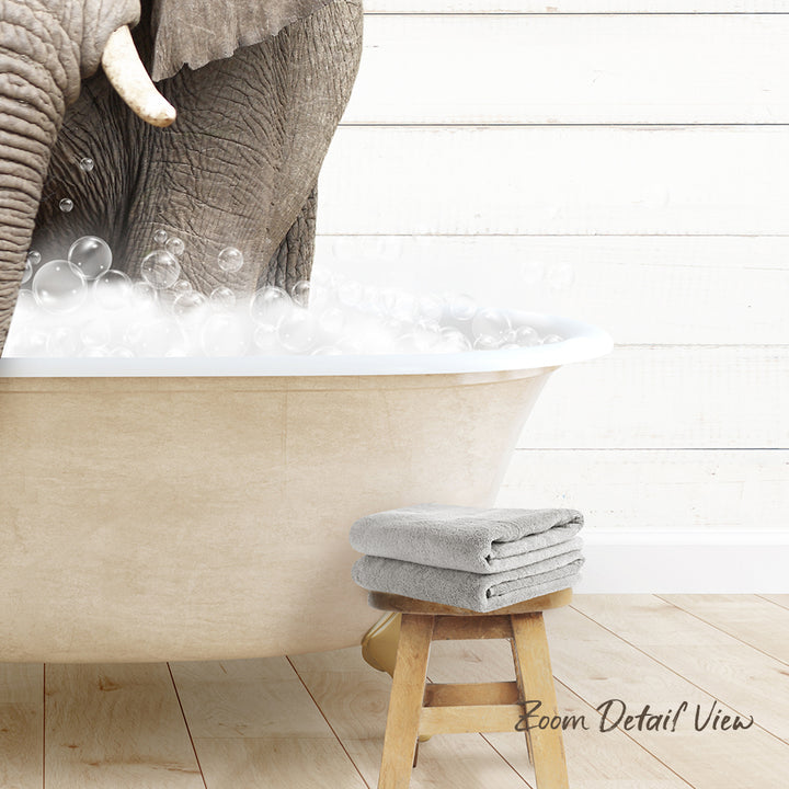 A large elephant is taking a bath in a bathtub filled with bubbles, with a towel and stool nearby.