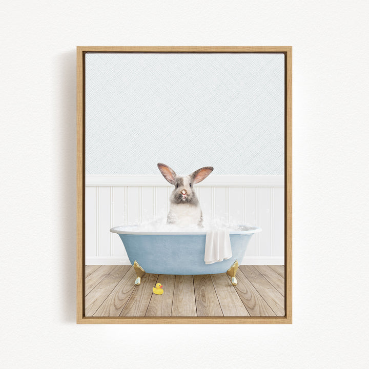 A gray and white rabbit sitting in a blue bathtub filled with bubbles, with a yellow rubber duck floating nearby.
