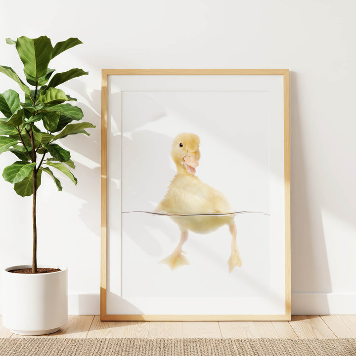 A framed print of a cute yellow duck swimming in water is displayed on a white wall, accompanied by a potted plant to the left.