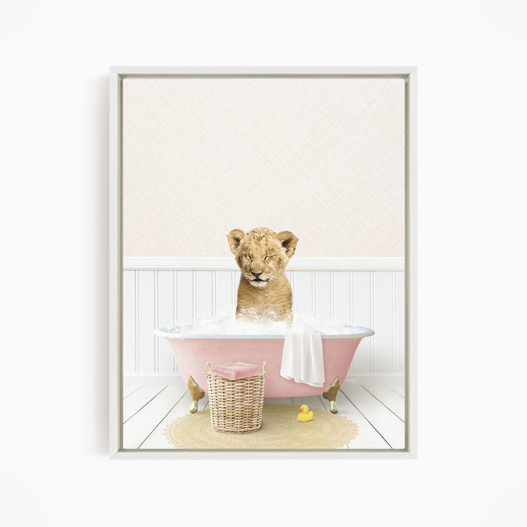 A young lion cub sitting in a pink bathtub filled with bubbles, surrounded by a yellow rubber duck and a basket.
