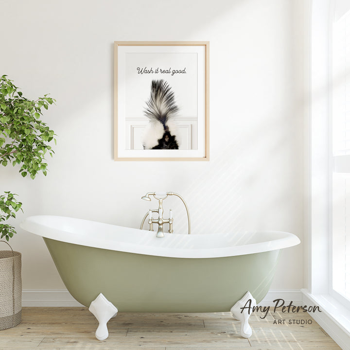 A framed print of a skunk's tail hangs on a white wall above a green claw-foot bathtub in a bright, airy bathroom.