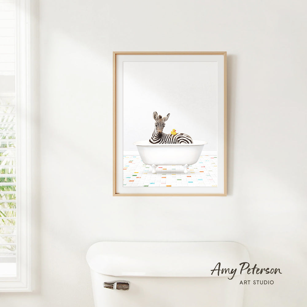 A framed print of a zebra sitting in a bathtub, surrounded by colorful tiles, is hanging on a white wall in a bathroom.