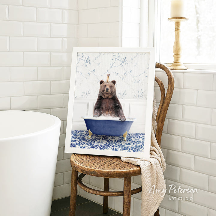 A framed image of a bear sitting in a blue bathtub, surrounded by a floral pattern, is displayed on a wooden chair in a bathroom setting.