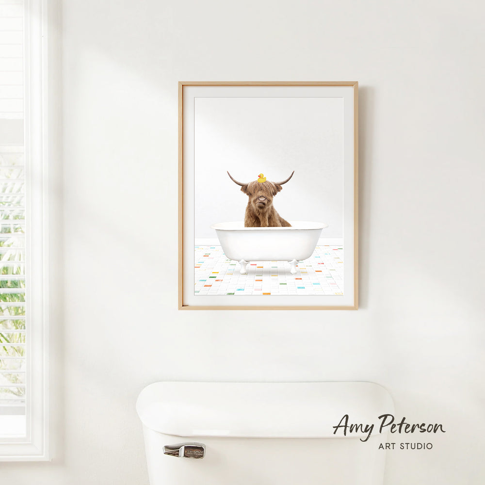 A framed image of a Highland cow sitting in a bathtub, with a yellow rubber duck on its head, is hanging on a white wall.