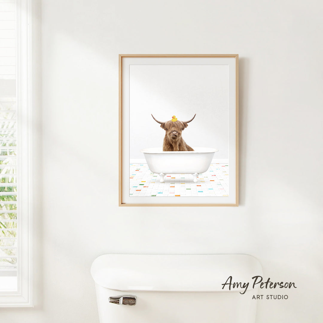 A framed image of a Highland cow sitting in a bathtub, with a yellow rubber duck on its head, is hanging on a white wall.
