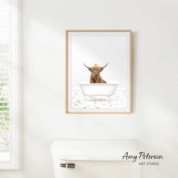 A framed image of a Highland cow sitting in a bathtub, with a yellow rubber duck on its head, is hanging on a white wall.