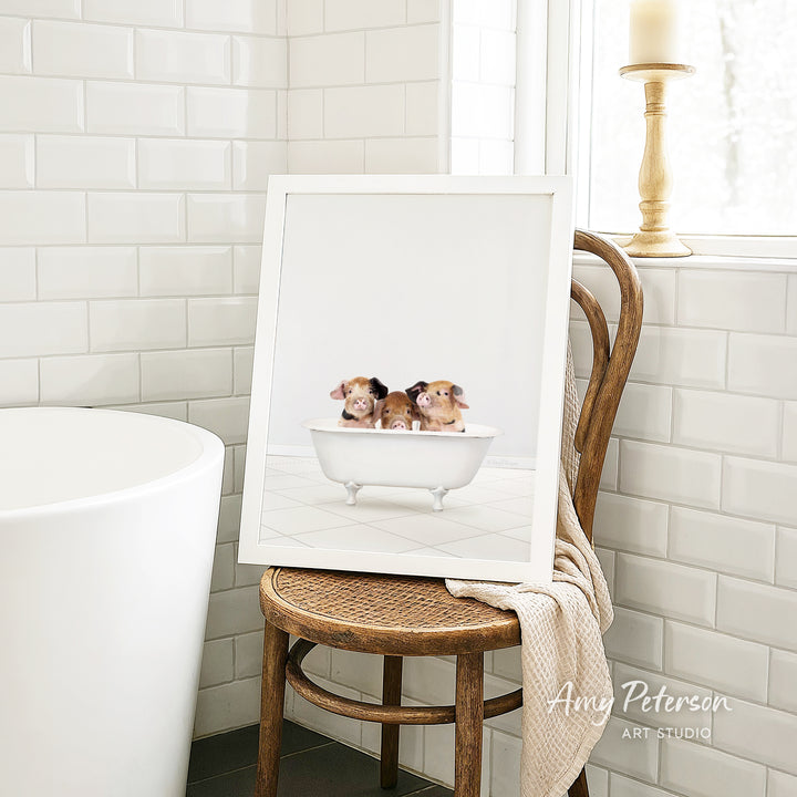 A framed image of three adorable pigs sitting in a bathtub is displayed on a wooden chair in a bathroom setting.