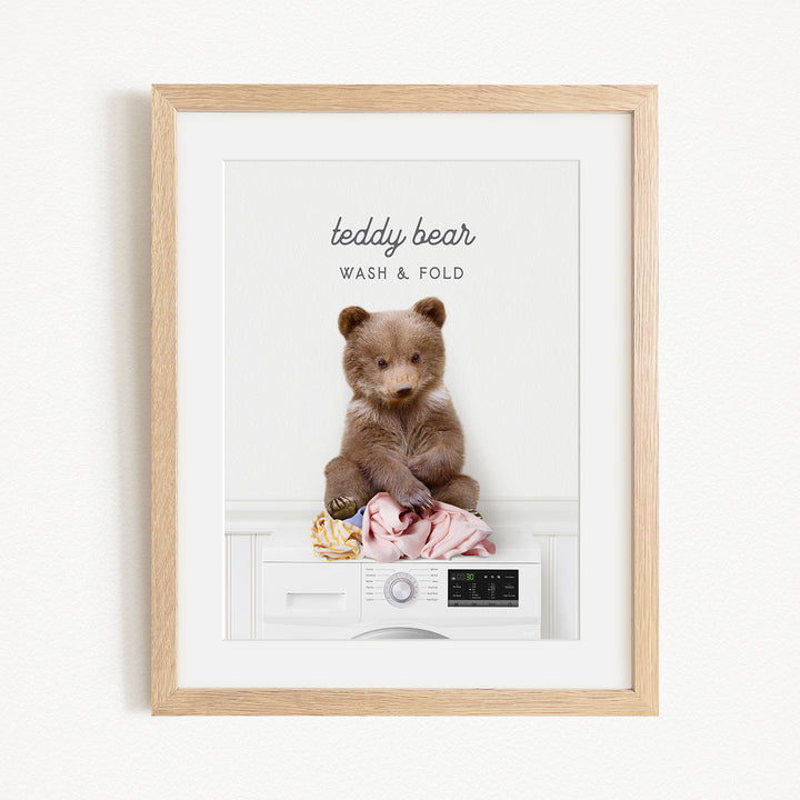 A teddy bear sitting on a washing machine, with clothes around it, and the text "teddy bear" and "WASH & FOLD" written above the bear.